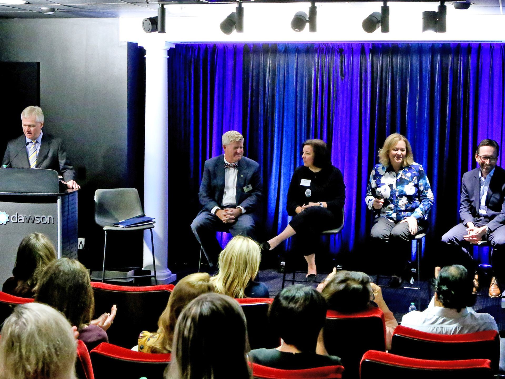 A panel of four professionals sits on stage before an audience, with a speaker at a podium to the left in a lit venue.