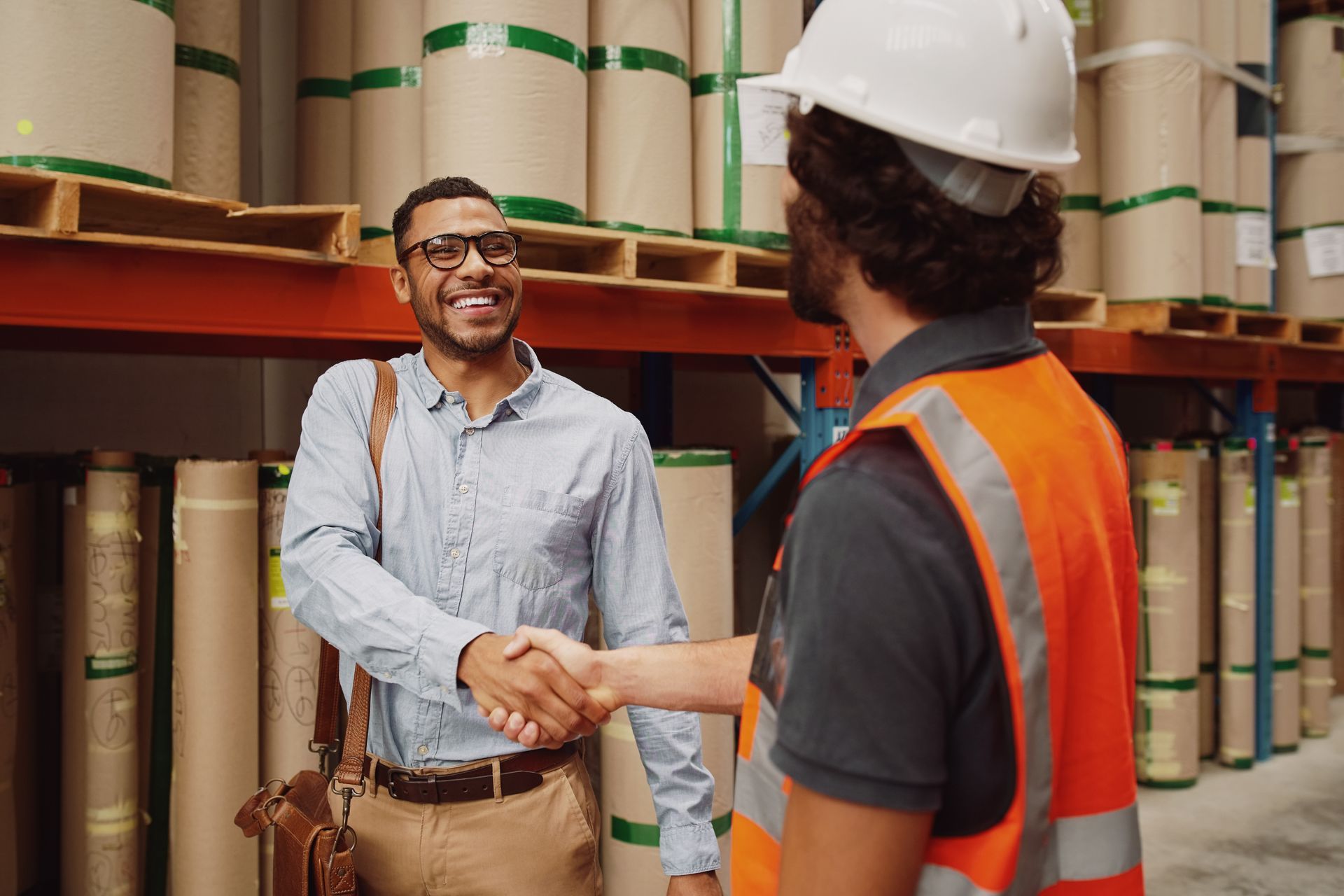 Two people shake hands in a warehouse aisle lined with stacked cardboard rolls, one wearing a safety vest and hard hat.