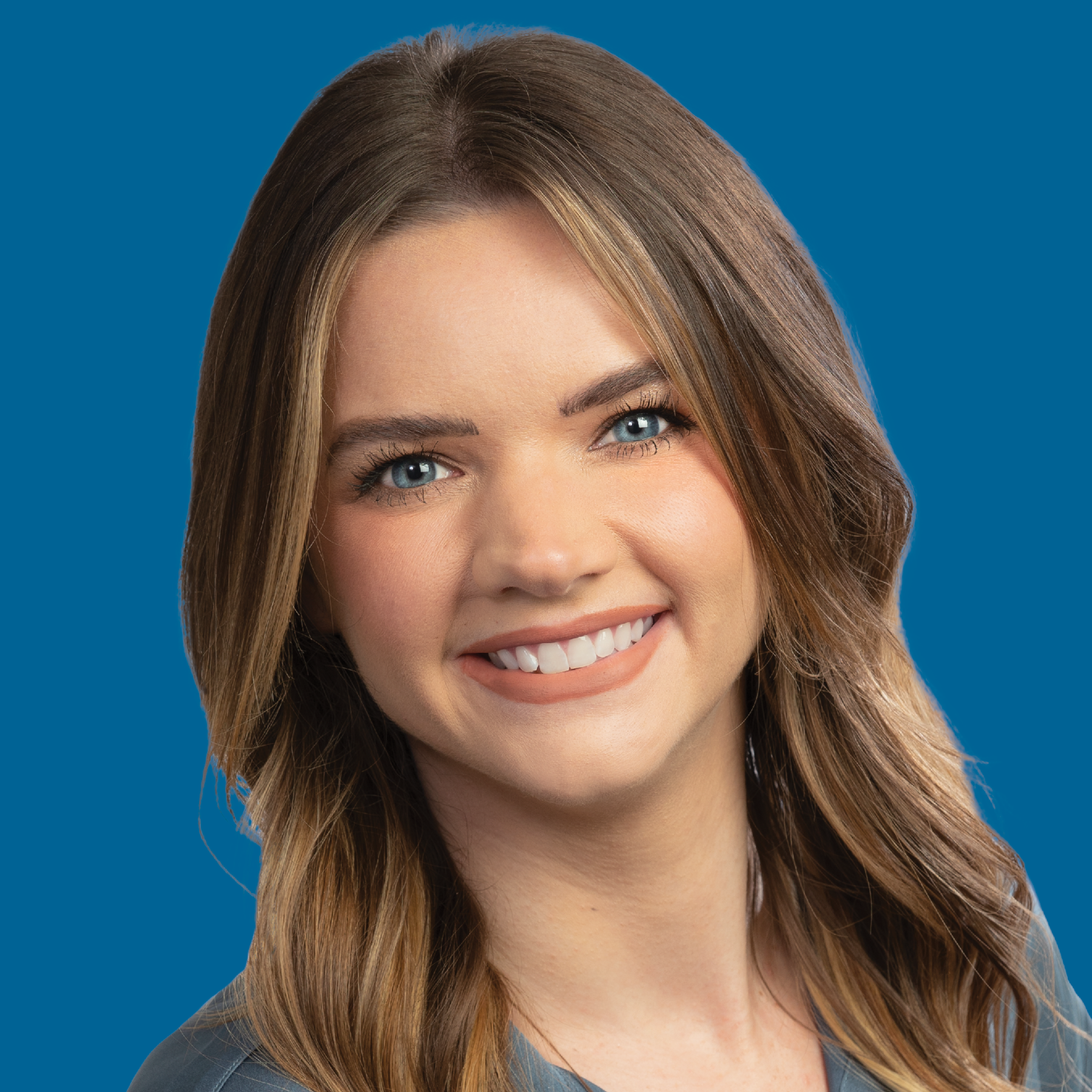 A professional headshot of a person with long brown hair, smiling against a solid blue background.