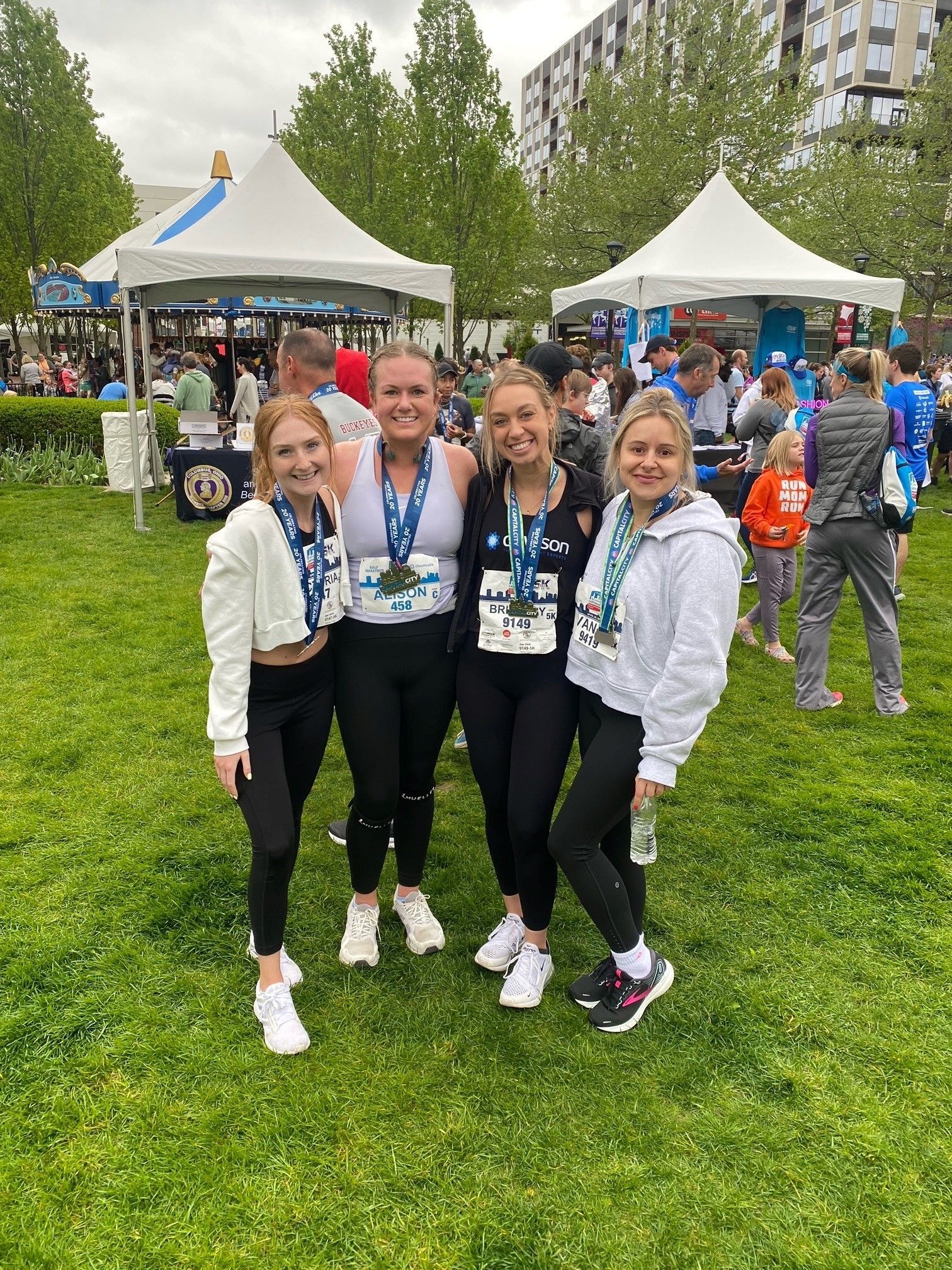 Four people wearing race bibs and medals pose together on a grass lawn near outdoor event tents.