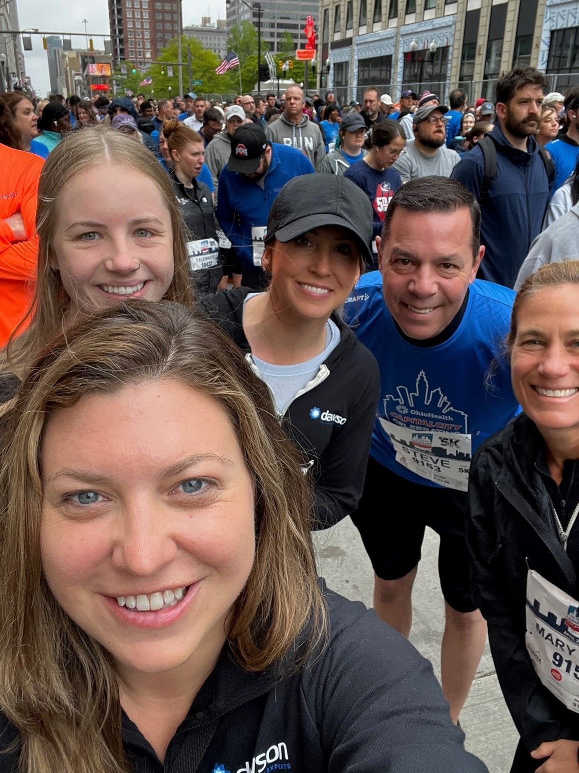 A smiling group takes a selfie at an outdoor race event, with other participants and buildings in the background.