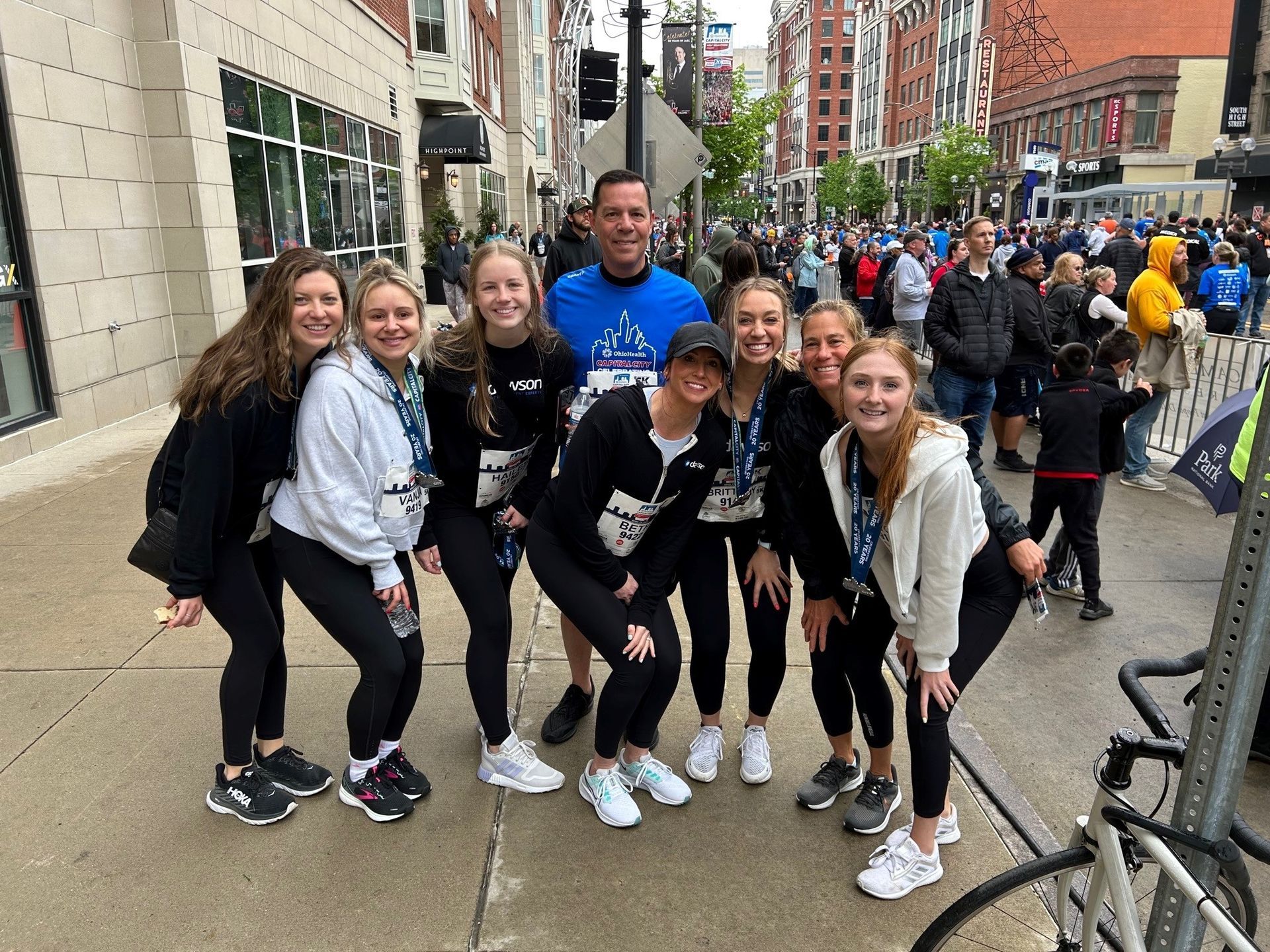 A group of people wearing matching black shirts and medals pose for a photo on a city sidewalk during a race event.