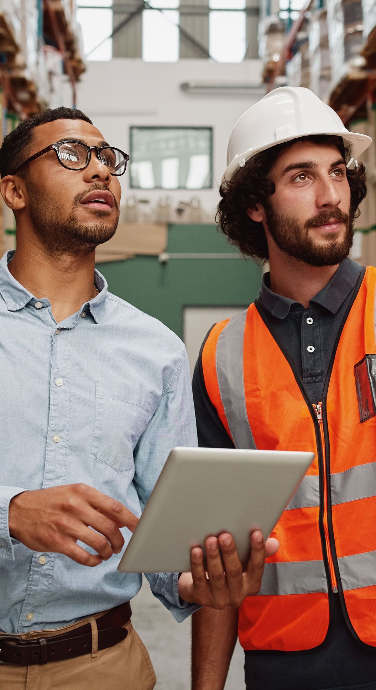 Two people in a warehouse, one holding a tablet and the other wearing a safety vest and hard hat, looking upward.