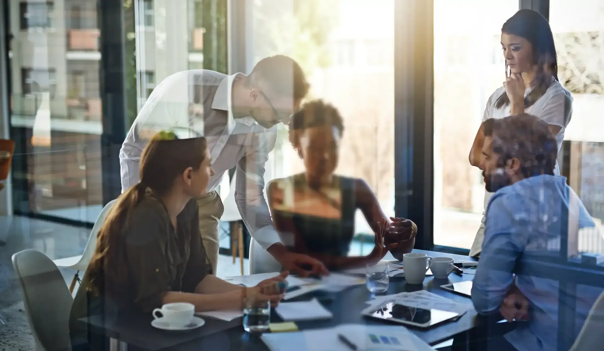 A diverse group of colleagues collaborates at a table in a modern office with glass walls and bright natural light.