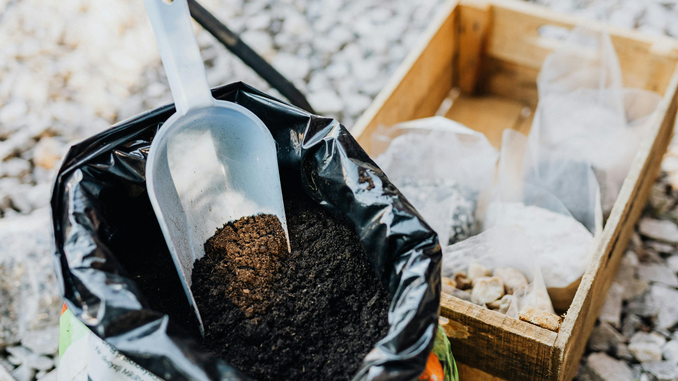 Black bag with dark soil, scoop inside. Wooden crate with sealed bags of material nearby, outdoor setting.