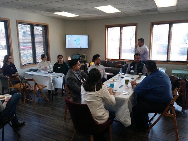 People seated around tables in a bright meeting room with a presentation screen.