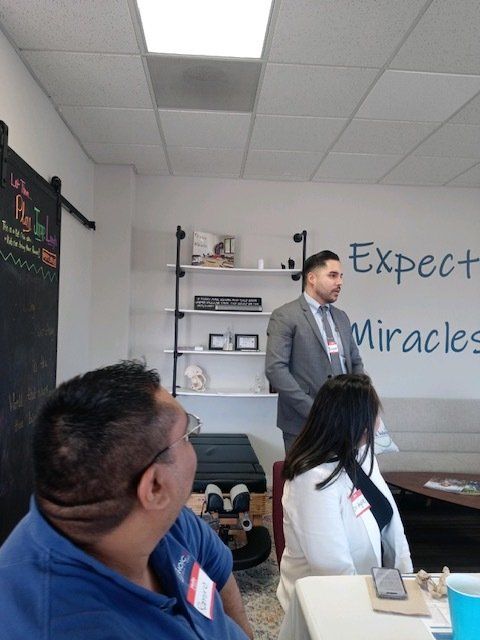 Man in gray suit speaks in a small classroom with “Expect Miracles” on the wall.