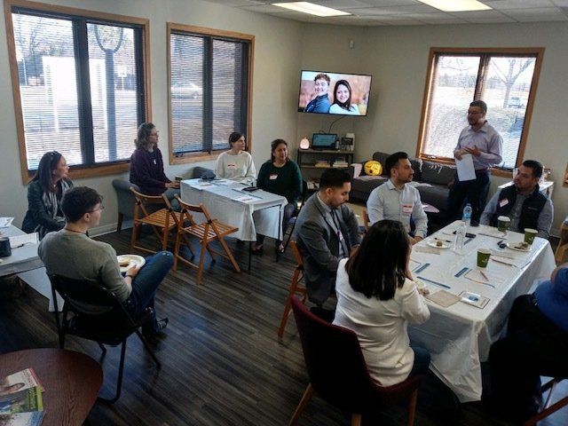Small meeting room with people seated around tables, laptops and papers out, during a group discussion on a video call.