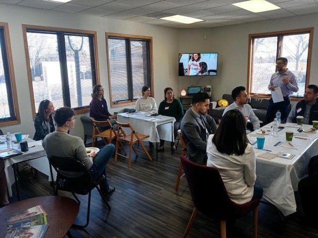 People seated at tables in a bright meeting room, listening to a wall-mounted TV during a group discussion.