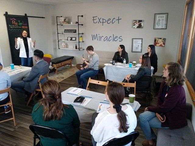 People seated around tables in a classroom meeting, with a presenter near a chalkboard reading “Expect Miracles.”