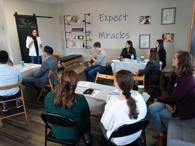 Small classroom workshop with people seated at desks while a presenter speaks beside a wall reading “Expect Miracles.”