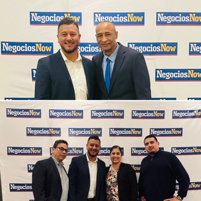Group of five people posing at a Negocios Now event backdrop, two rows, smiling in business attire