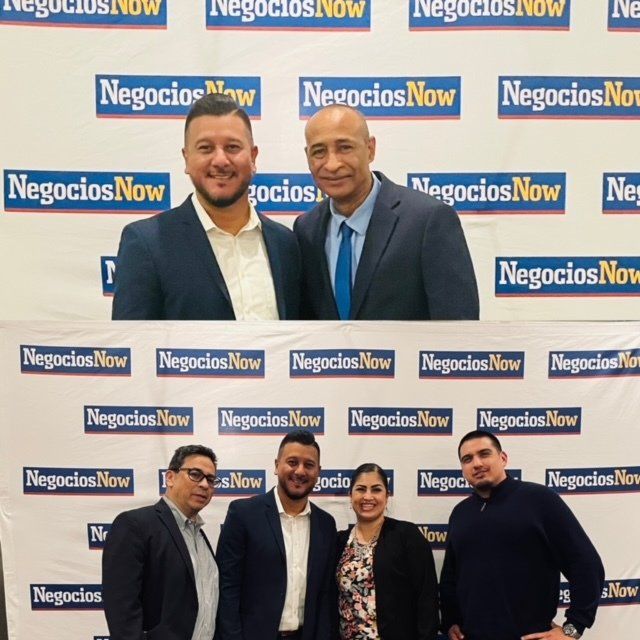 Five people posing in front of a NegociosNow backdrop at a networking event