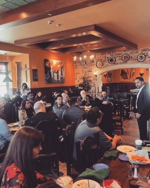 People seated in a cozy restaurant, listening to a man speaking near the front.
