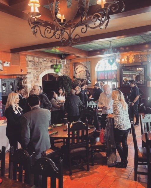 Crowded restaurant interior with people standing and chatting under chandeliers and warm lighting.