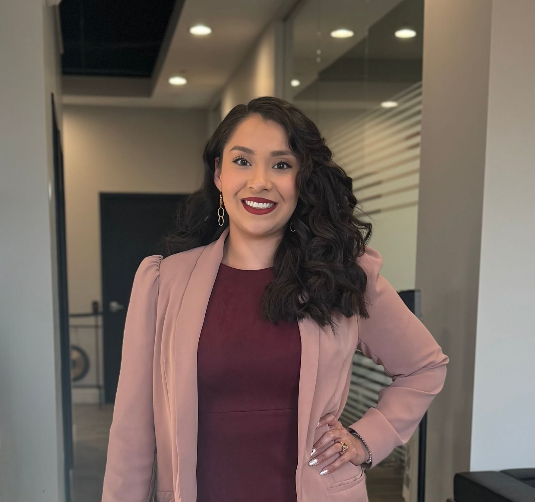 Woman smiling in a mauve blazer and burgundy dress, standing in a modern office hallway.