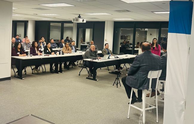 Audience seated at tables in a conference room, listening to a speaker at the front.