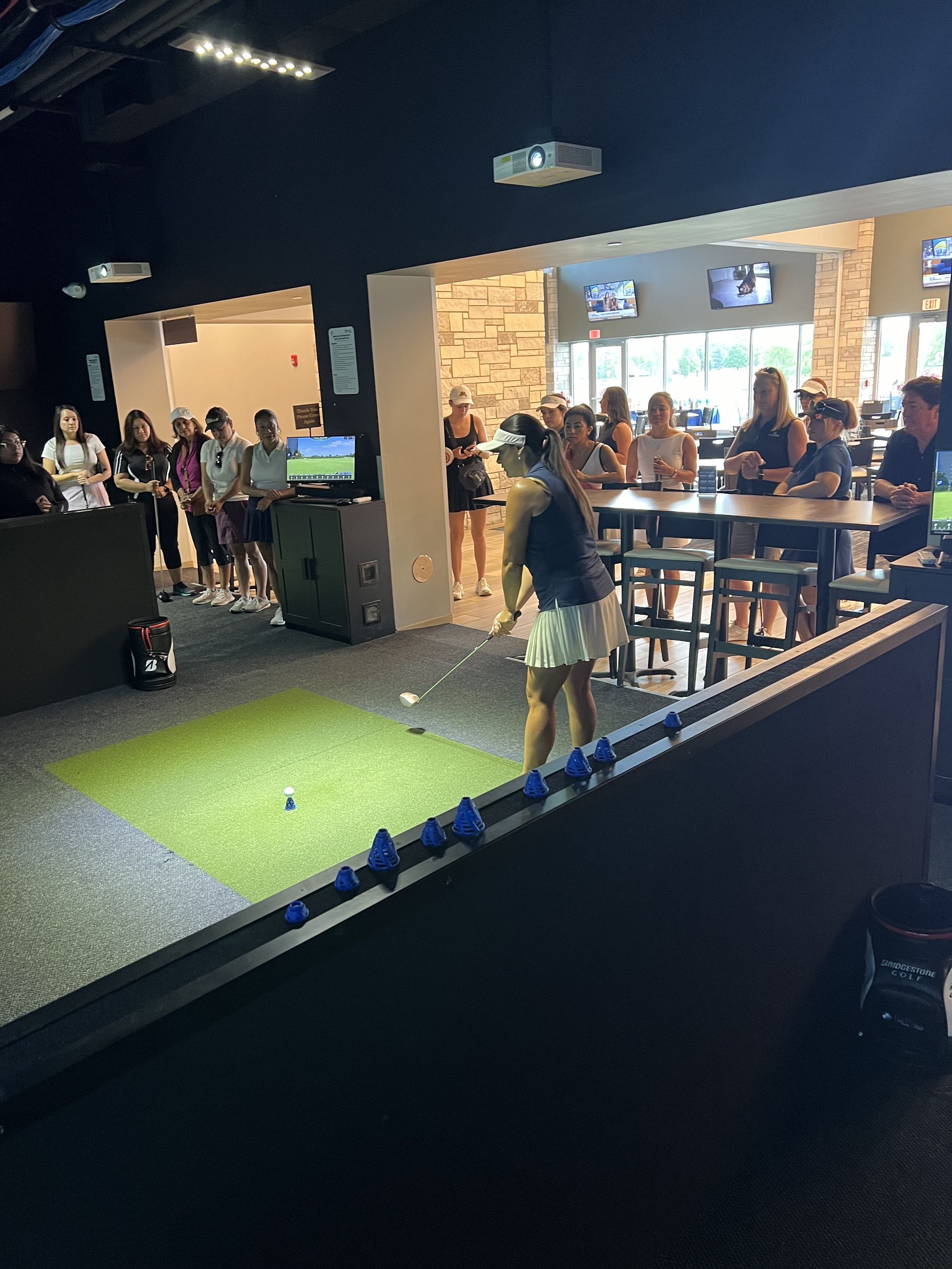 Indoor mini-golf scene with a person putting on a green turf lane while others watch in a dark, open venue.