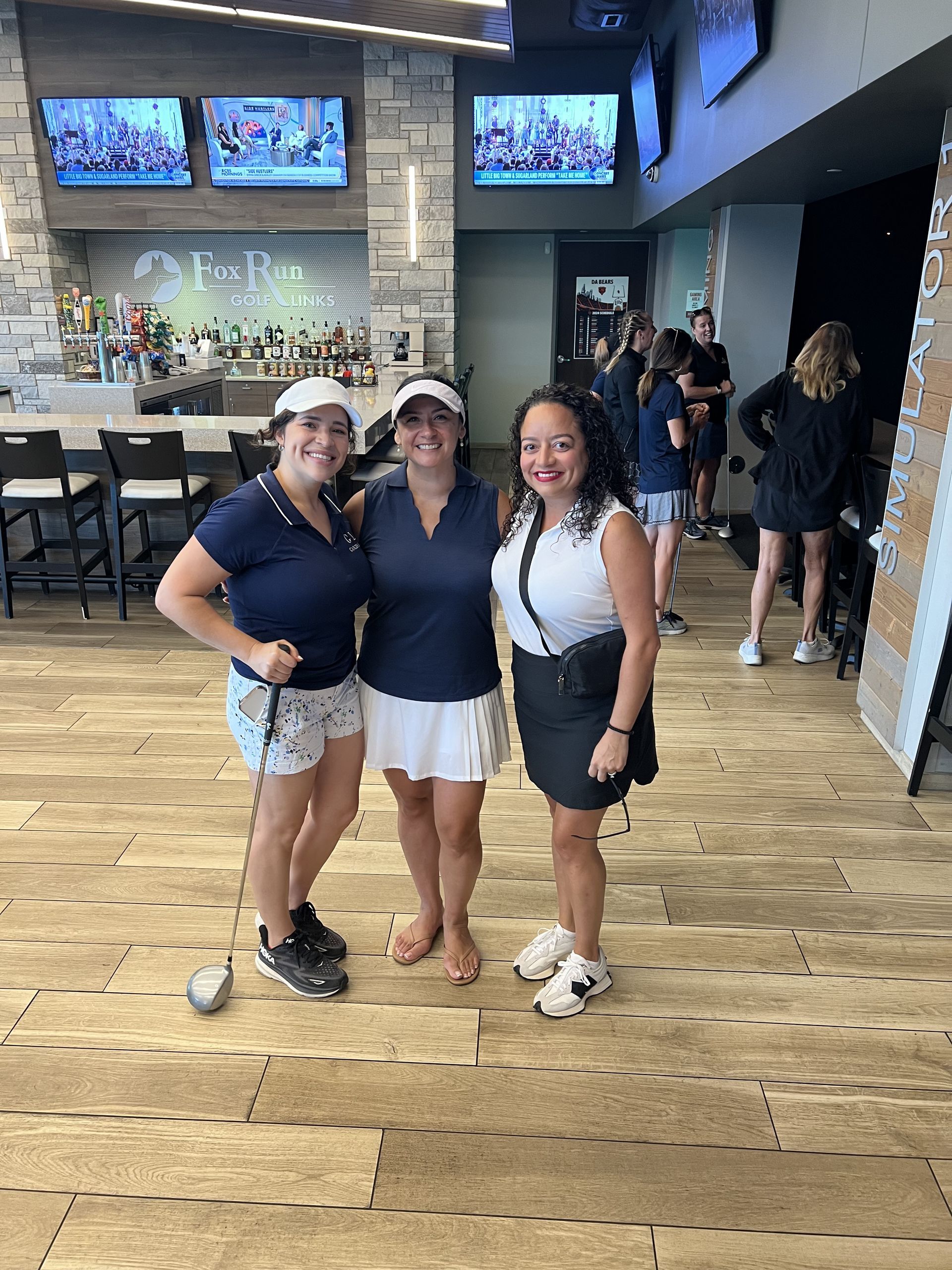 Three people smiling and posing in a bar with TVs and a wooden floor.