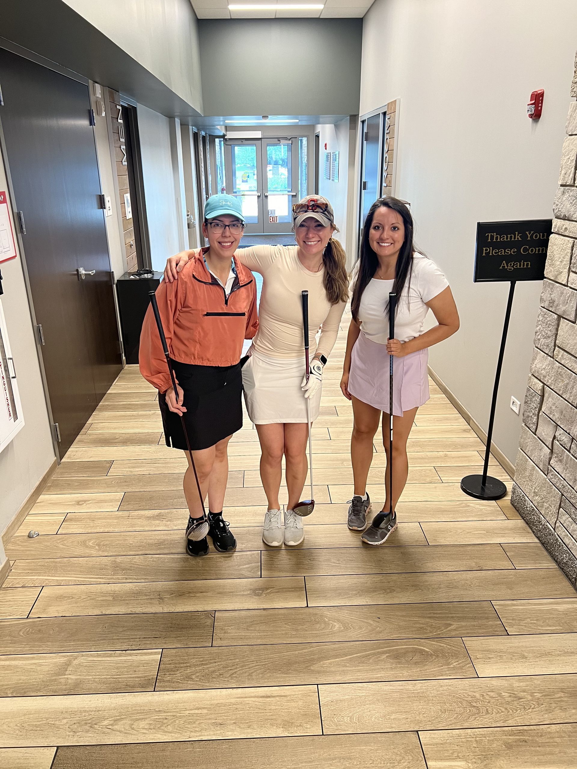 Three friends smiling in a hallway, standing arm-in-arm, with a beachy floor and gray walls.