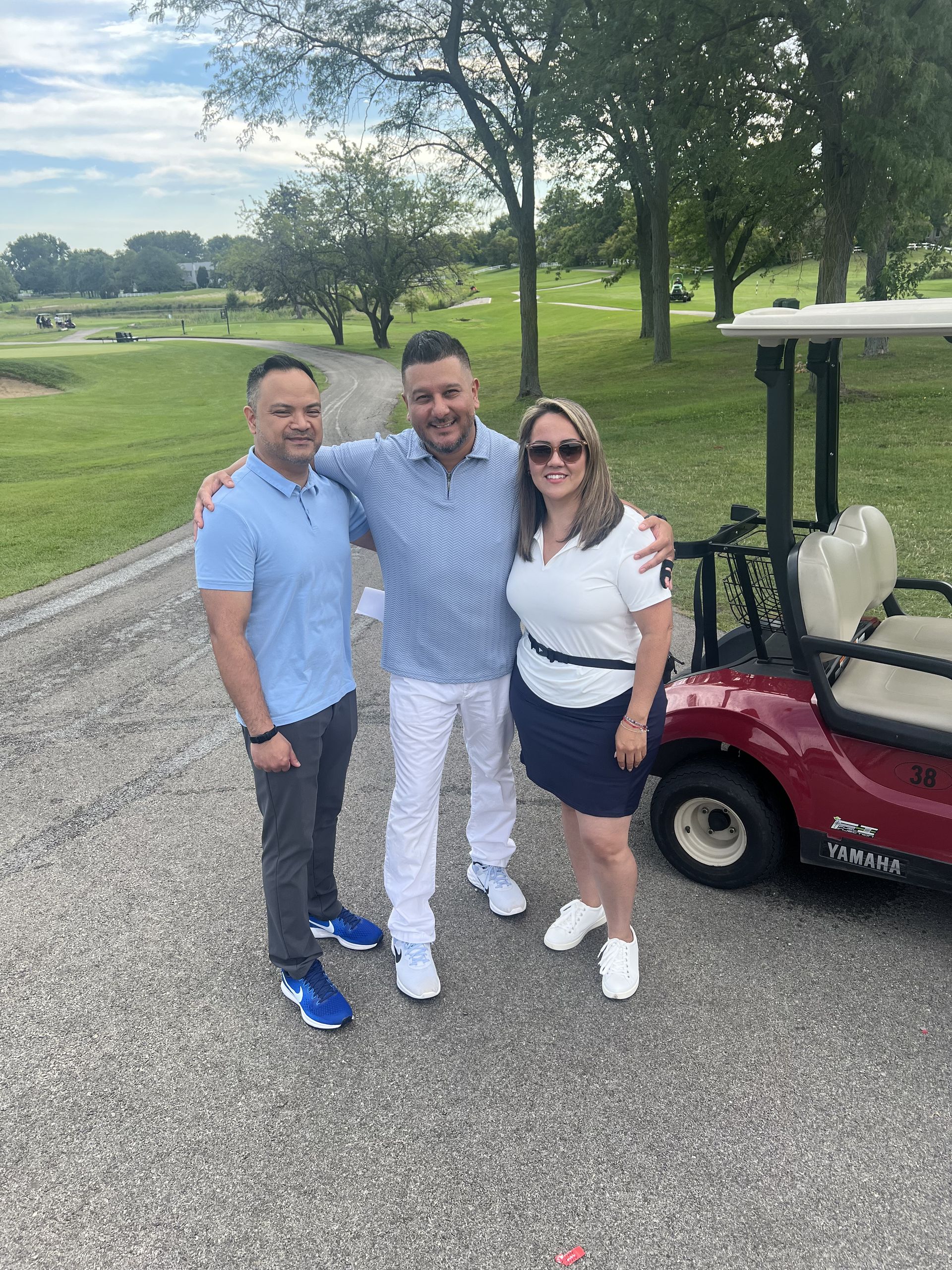 Three people posing together on a gravel path beside a golf cart in a grassy park.