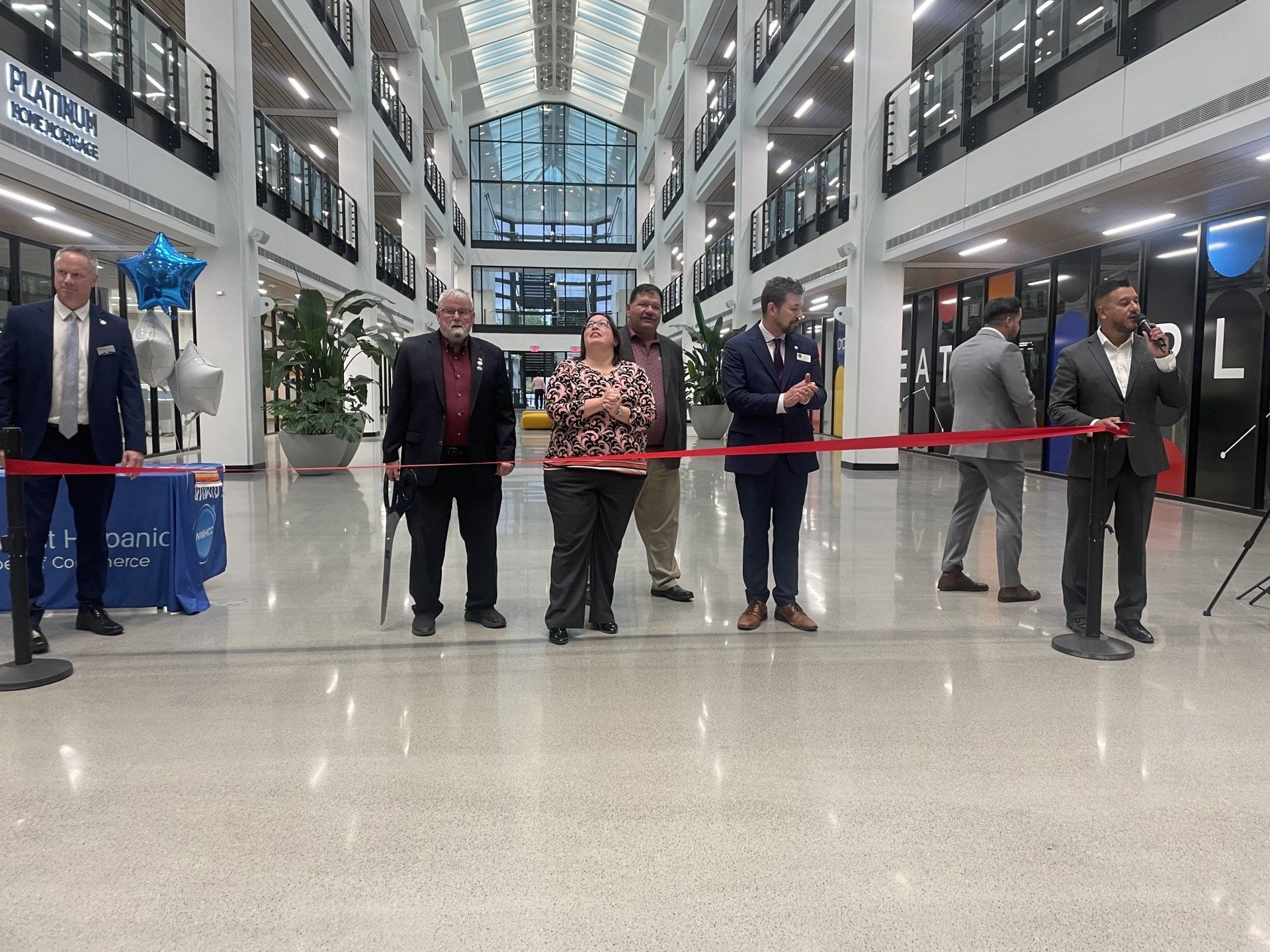 People standing behind a red ribbon in a bright indoor atrium, as if at a grand opening.