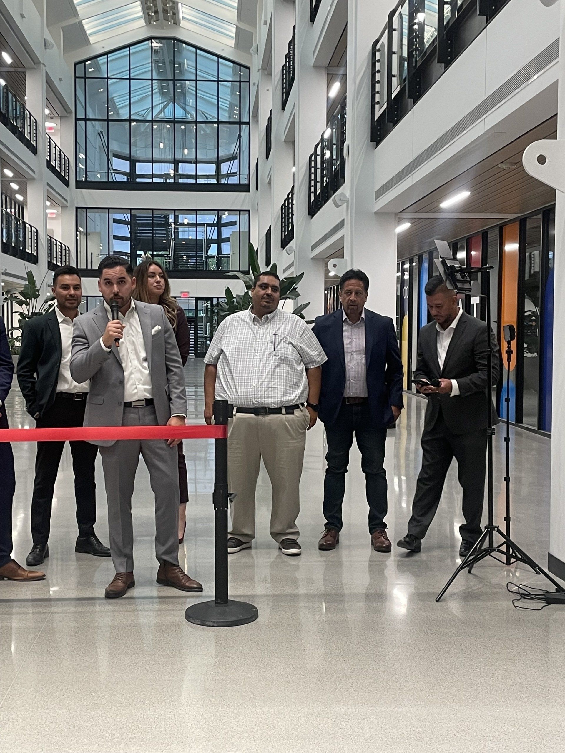 Six men stand behind a red ribbon in a bright atrium during a ribbon-cutting ceremony.
