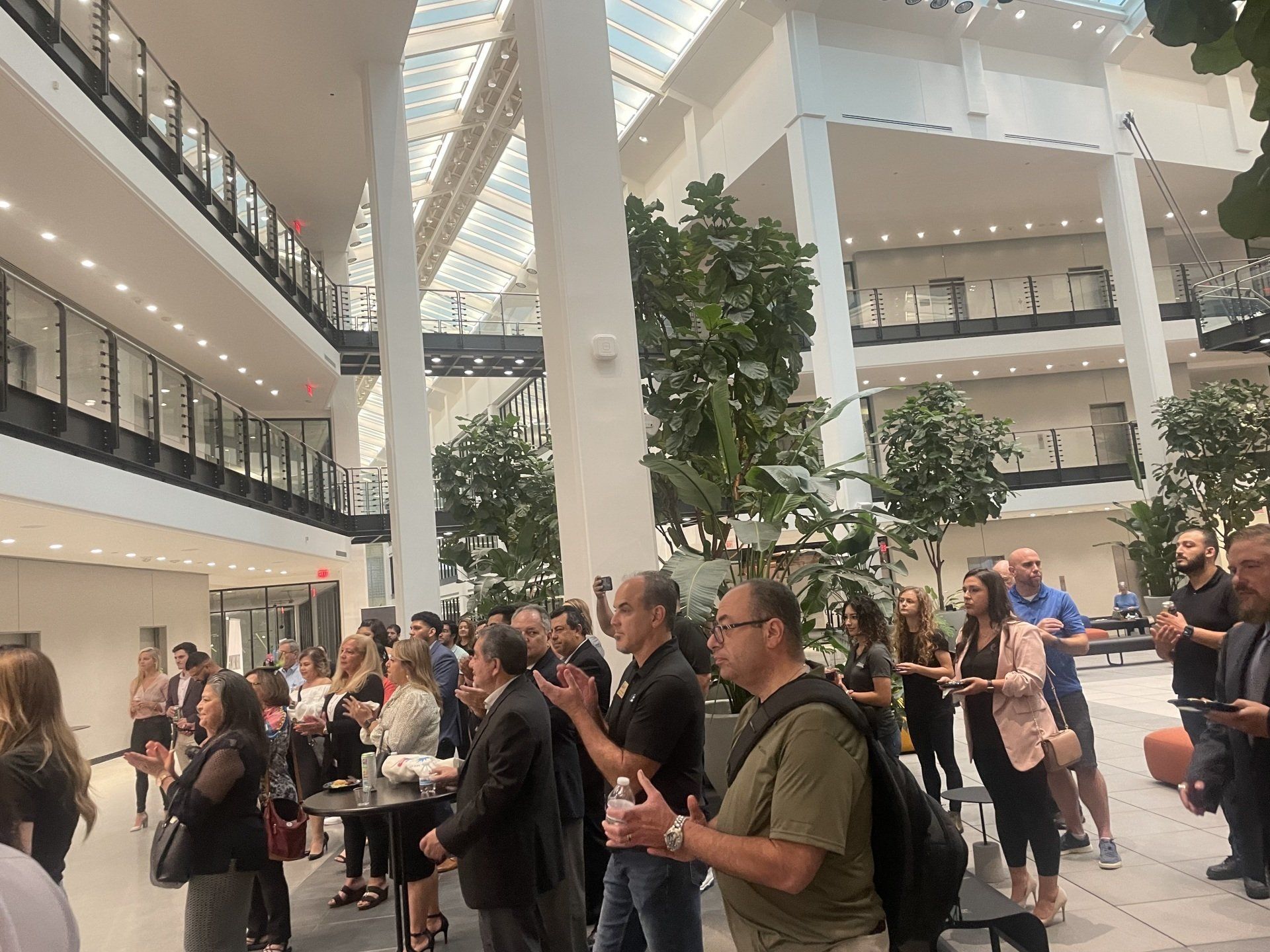 Crowd gathered in a bright atrium, listening and socializing near tables and tall indoor trees