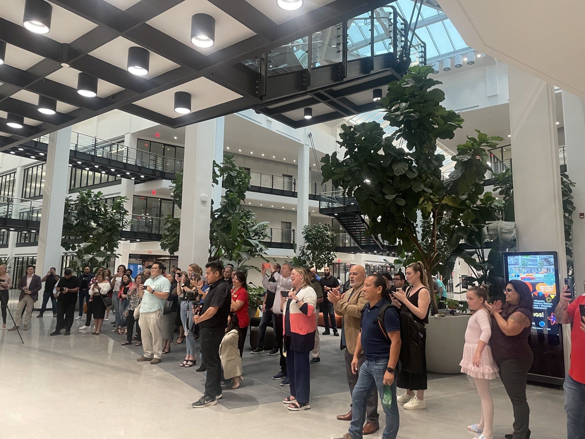 Crowd gathered in a bright indoor mall atrium beneath glass walkways and potted trees