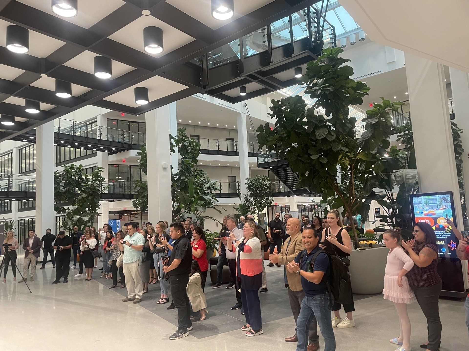 Crowd standing in a bright indoor atrium, waiting near tall plants and glass walkways