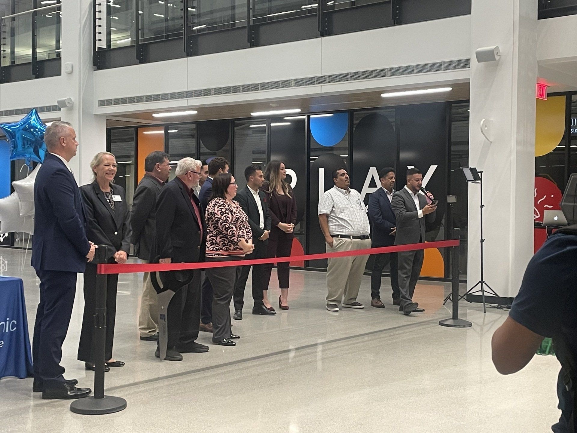 People gathered at a ribbon-cutting event in a bright lobby, with red ribbon and balloons.