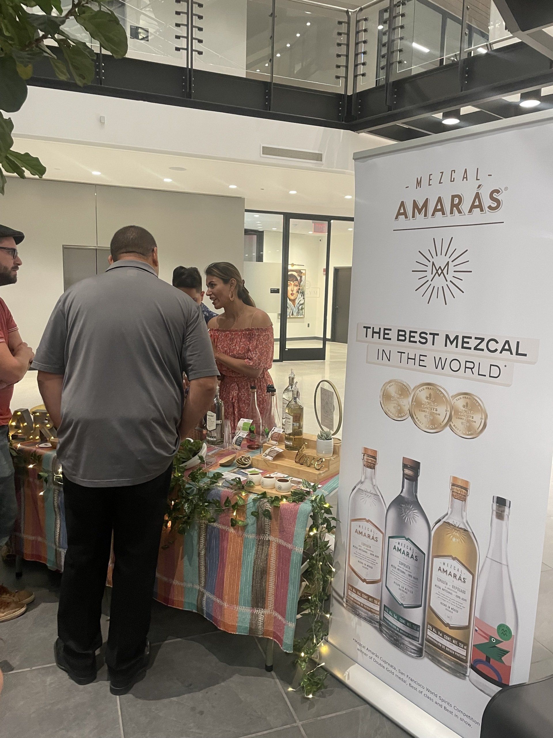 People at a mezcal tasting booth beside a colorful table and AMARAS banner in a bright indoor venue
