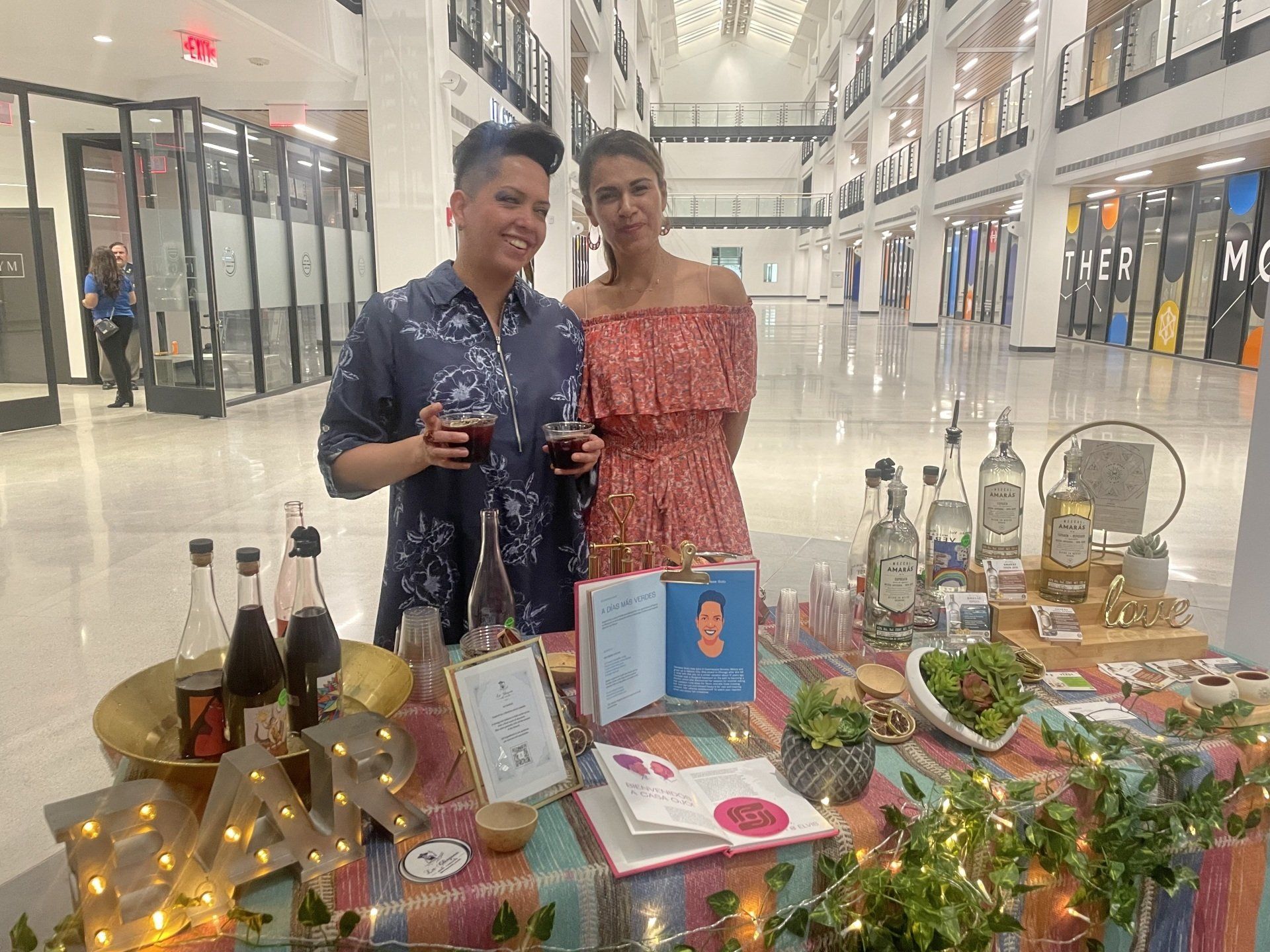 Two people posing beside a decorated table with candles, flowers, and bottles in a bright indoor hall