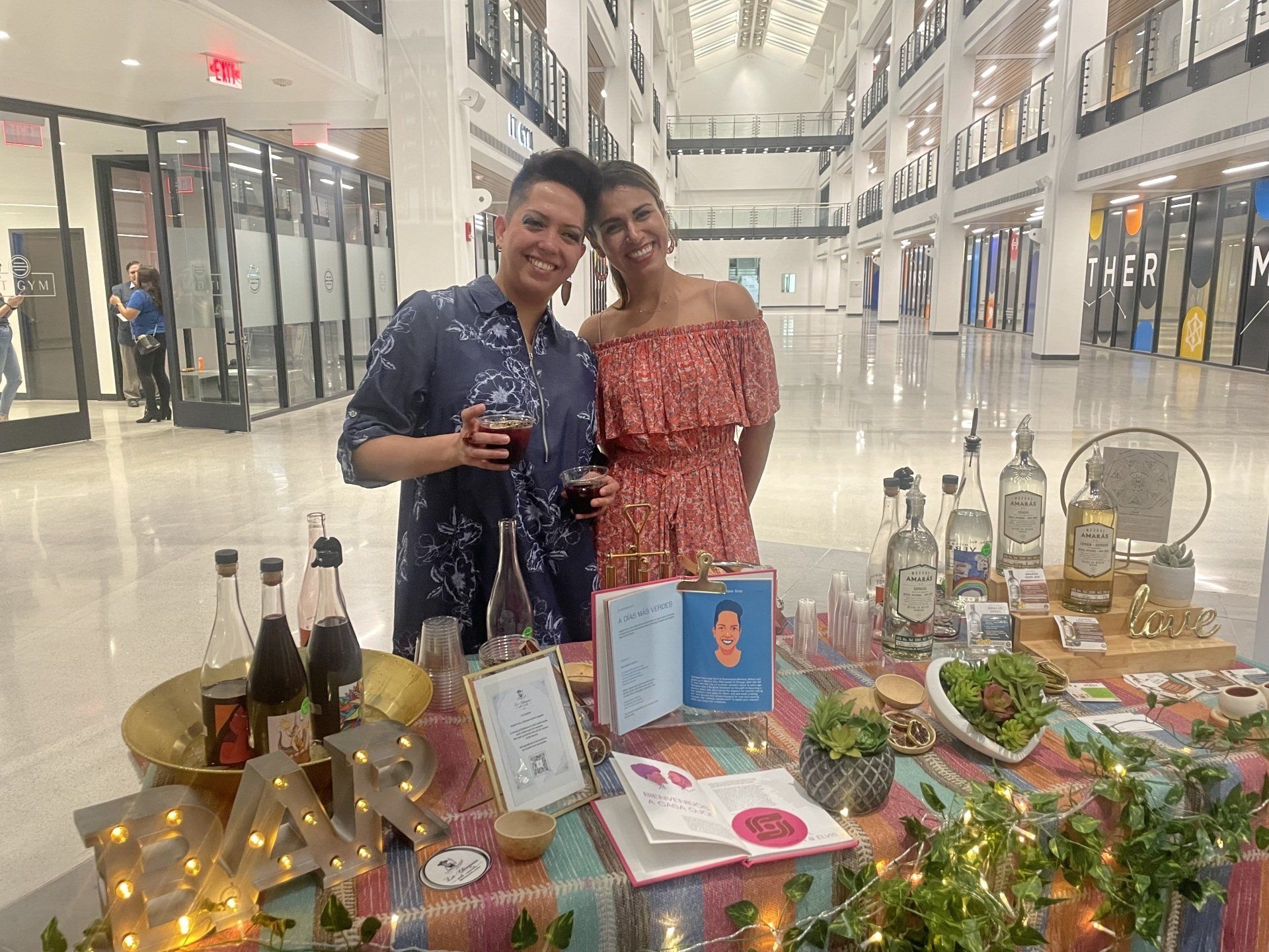 Two people posing behind a decorated table with bottles, flowers, and lights in a bright indoor mall.
