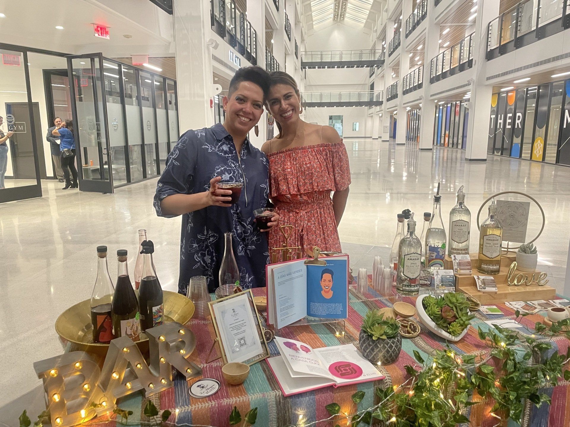 Two people pose behind a decorated table with bottles, flowers, and candles in a bright indoor mall.