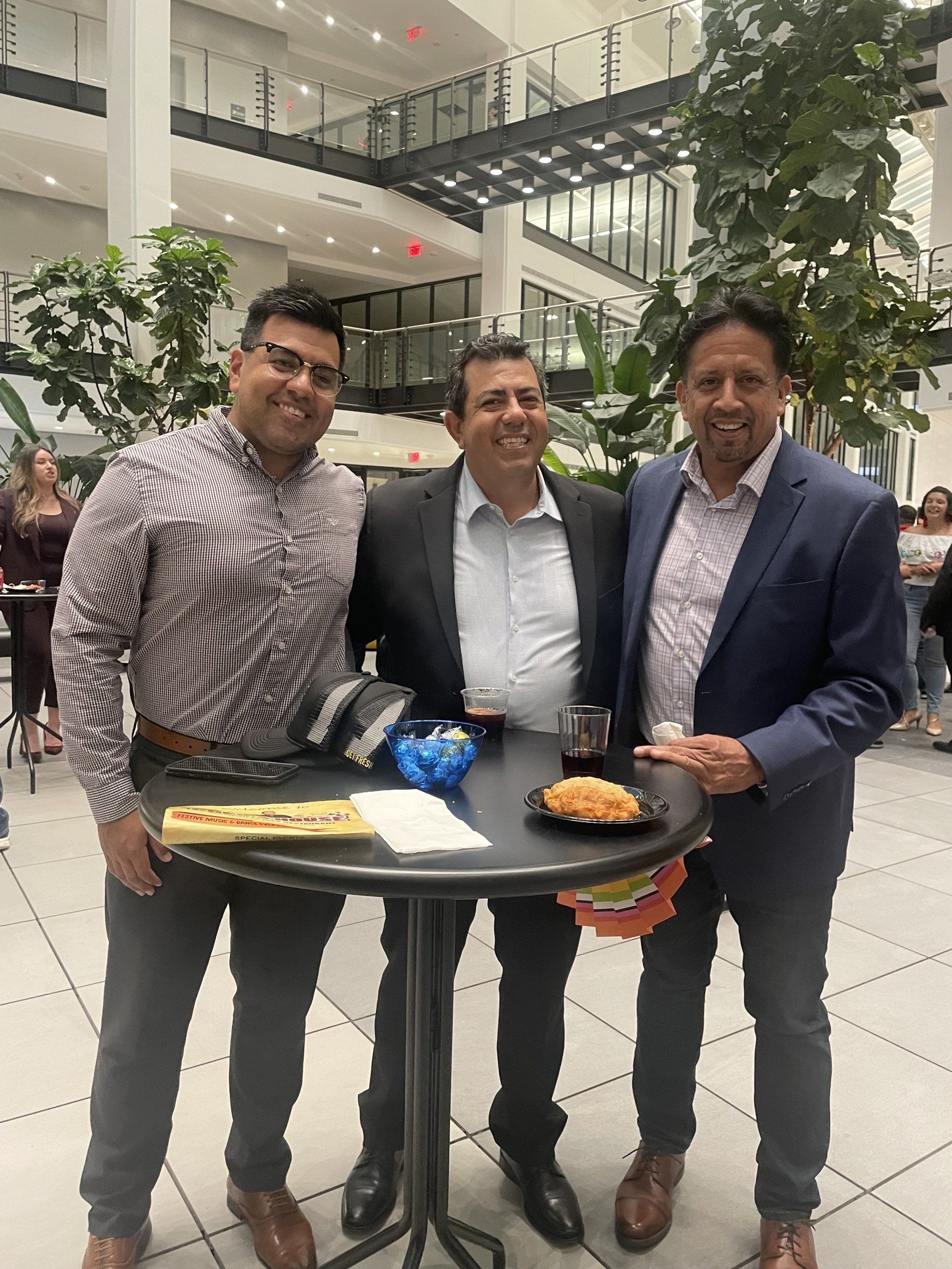 Three men in suits posing with drinks and snacks at a modern indoor atrium