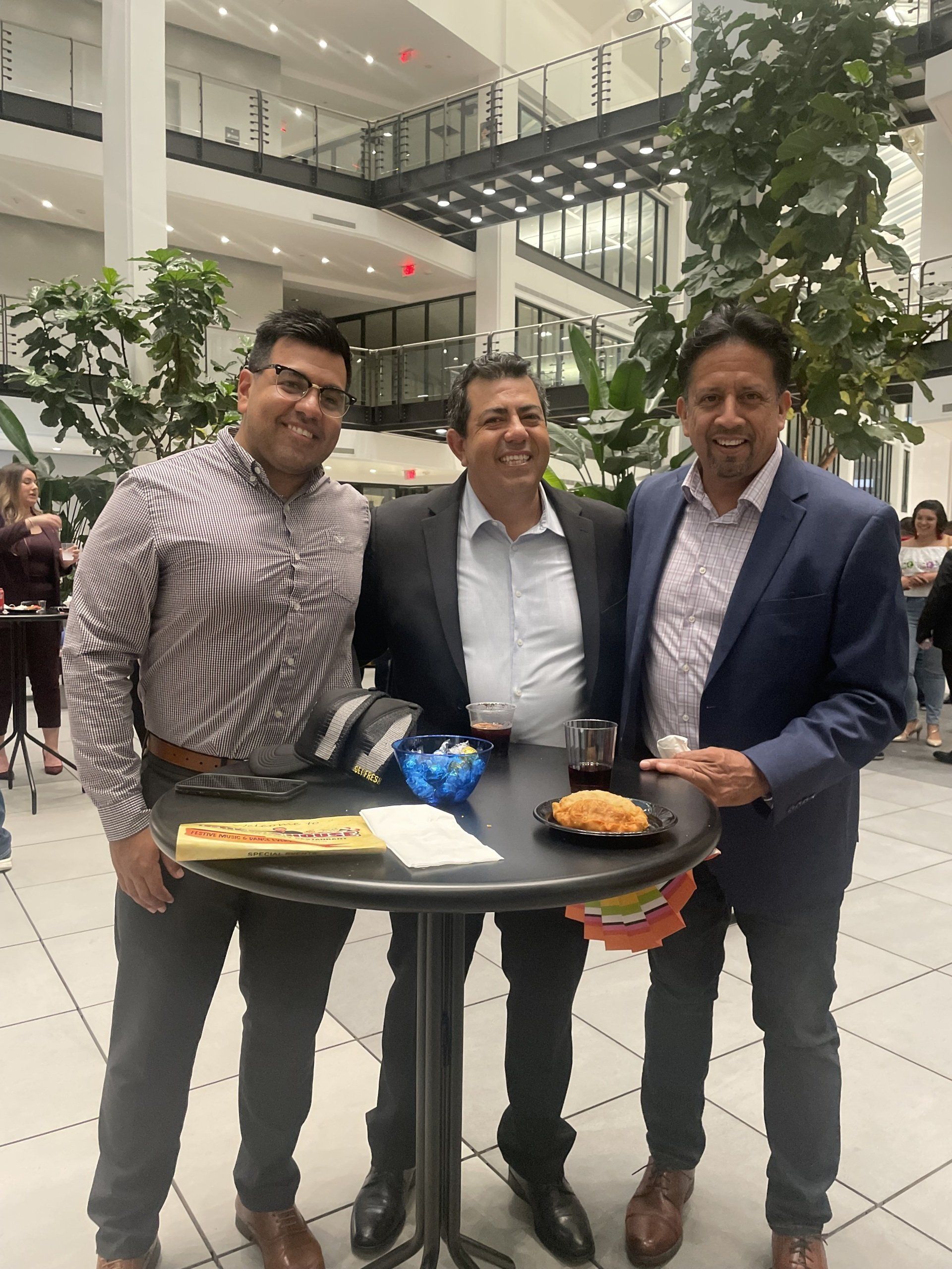 Three men in suits posing indoors with a tray of drinks and pastries in a bright atrium
