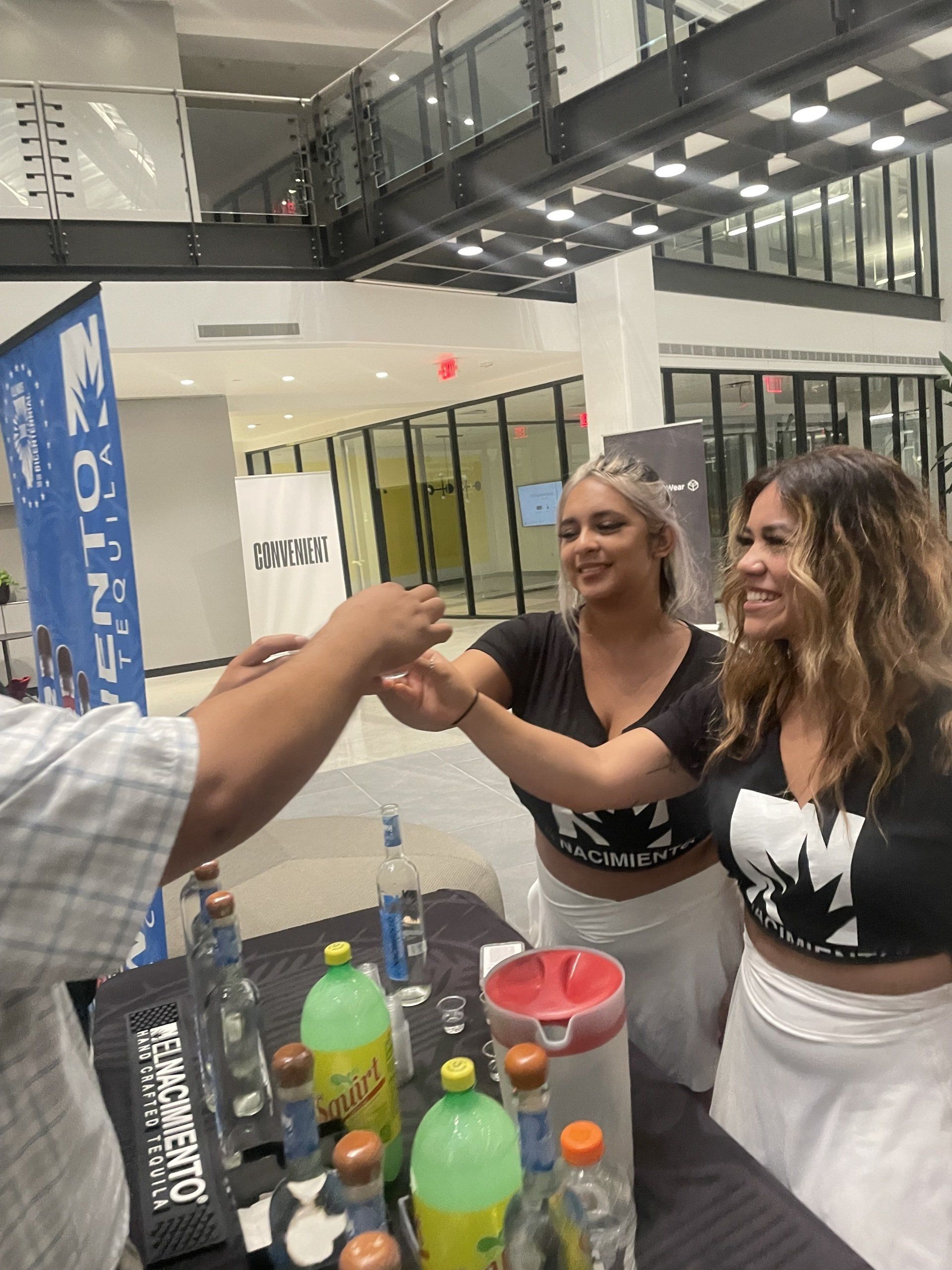 Two women behind a drink table smiling and reaching to toast with a guest in a bright indoor atrium.
