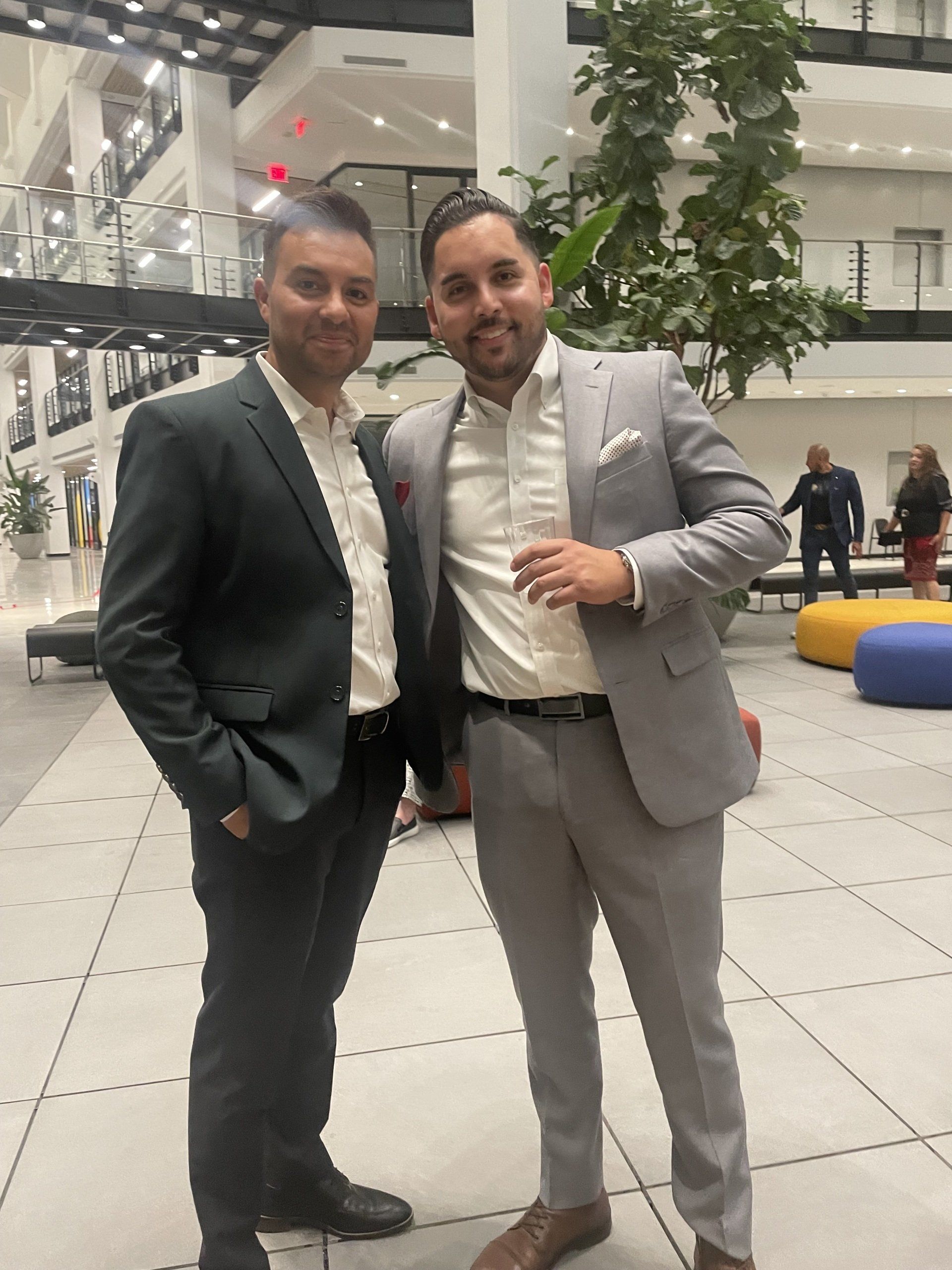 Two men in suits posing indoors in a modern lobby with plants and seating.
