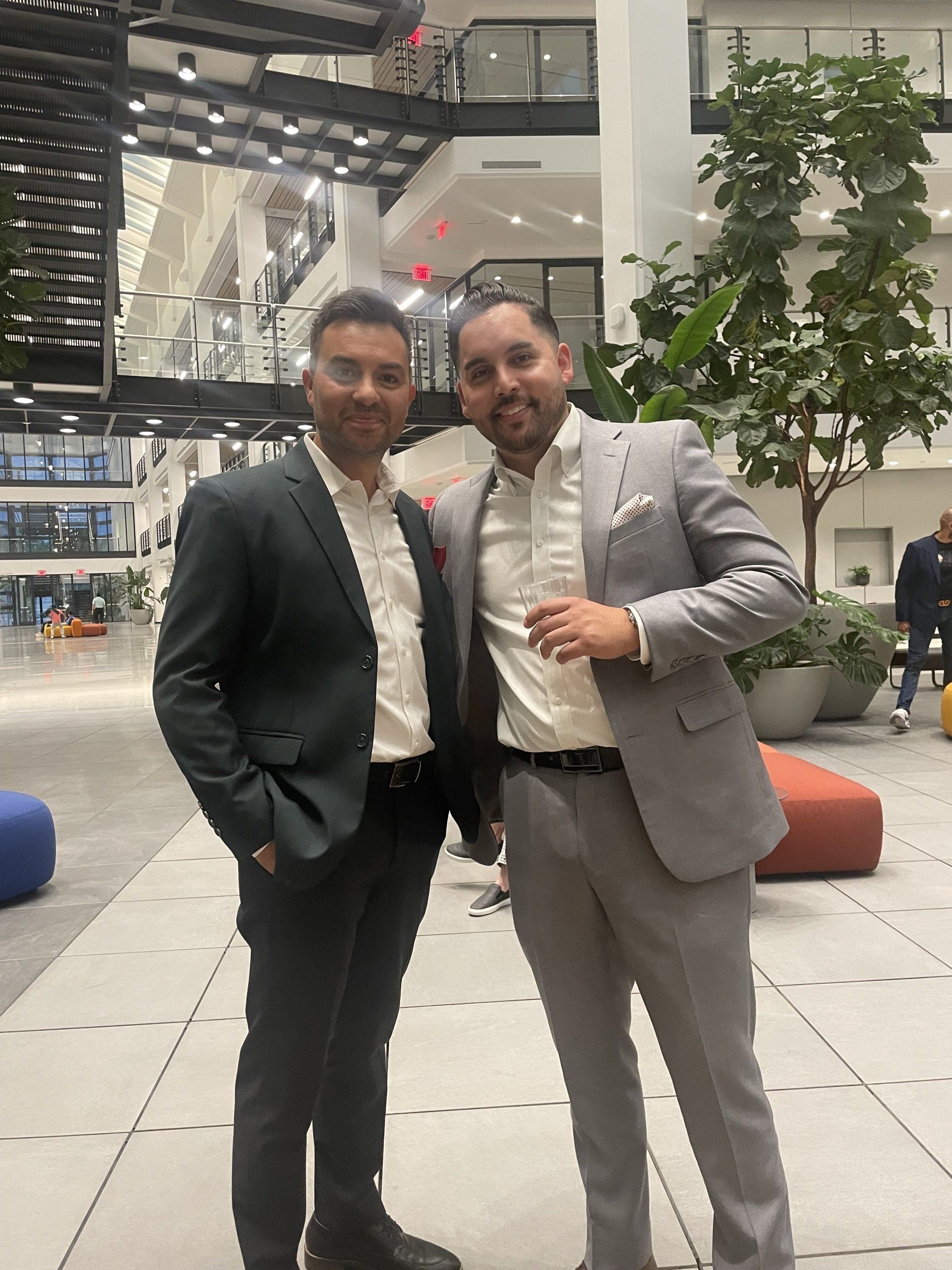 Two men standing together in a modern indoor lobby, both in suits and smiling at the camera.