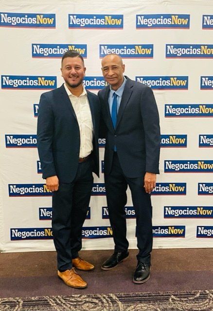 Two men in suits posing in front of a NegociosNow backdrop at a formal event