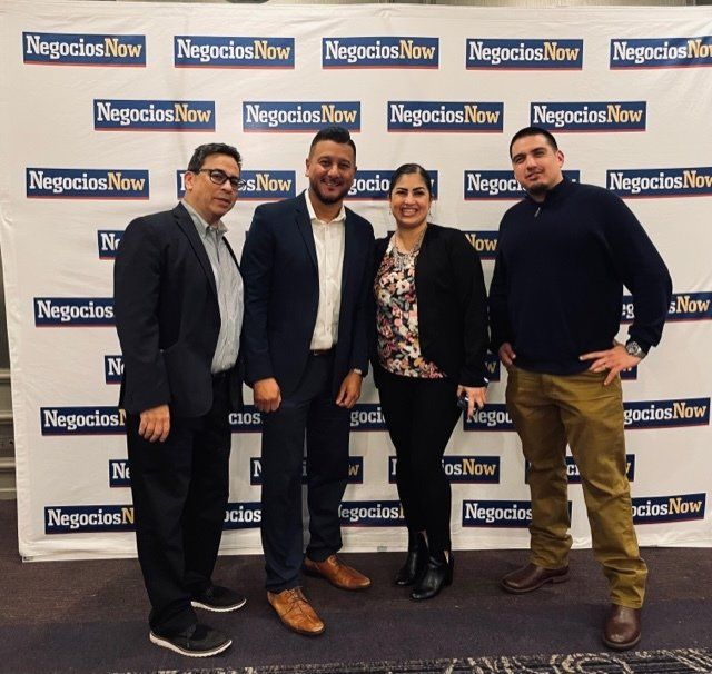Four people posing at a Negocios Now event backdrop, smiling and dressed in business attire.