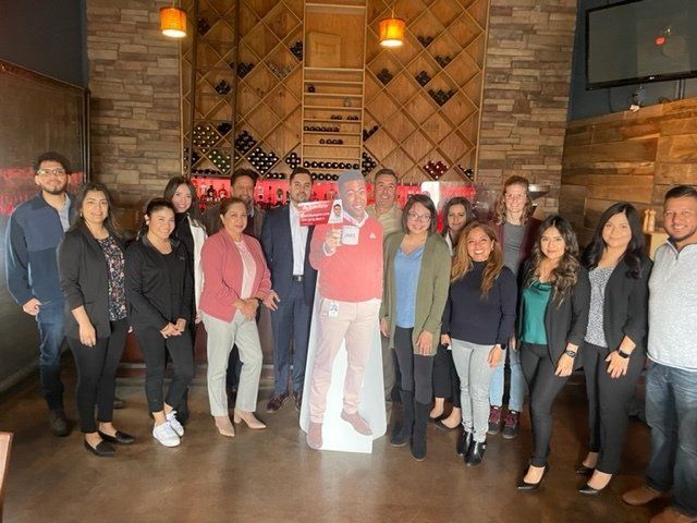 Group of people posing indoors in a rustic venue, standing in front of a patterned wall and lit sign.