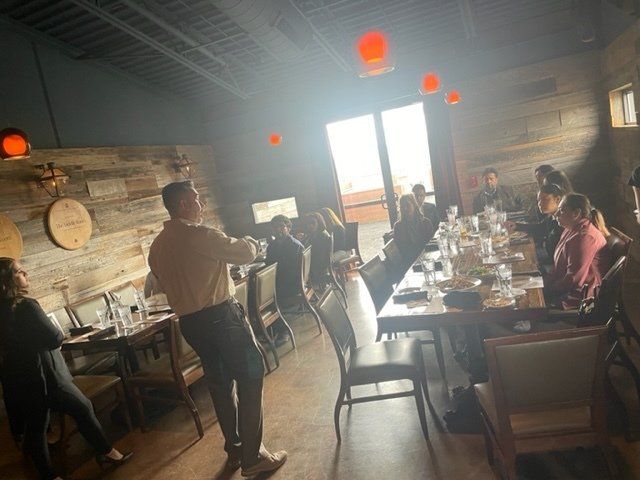 People seated at a long dining table in a rustic restaurant, with sunlight streaming through the open door.