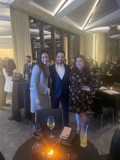 Three people posing at a dimly lit restaurant event with drinks and city lights behind them