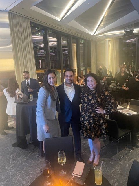 Three people posing at a formal indoor event with tables, drinks, and warm lighting