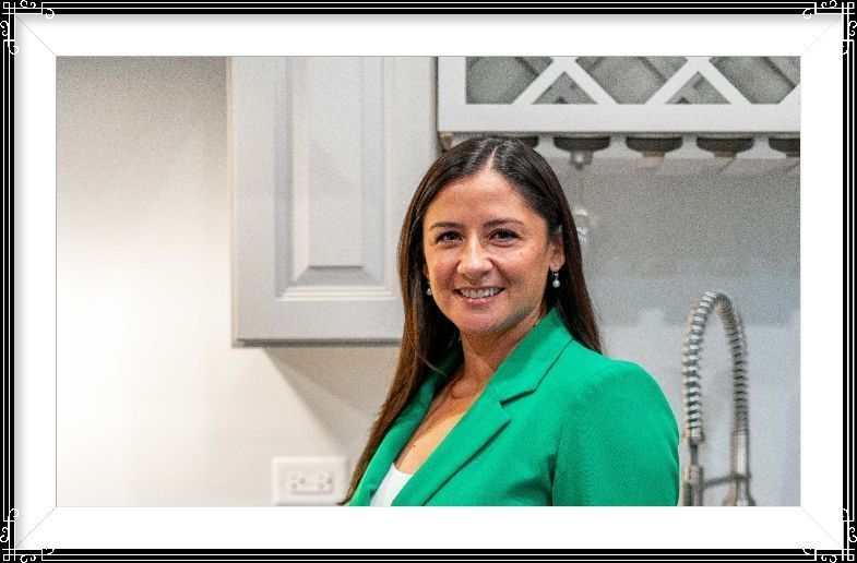 Woman in a green blazer smiling in a kitchen beside white cabinets and a sink