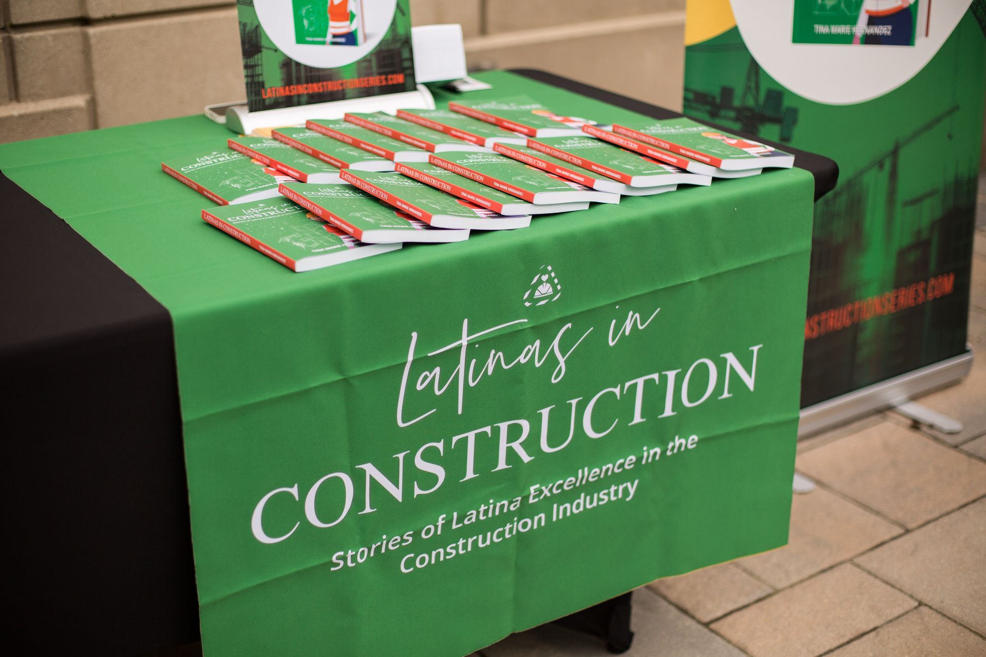 Green “Latinos in Construction” table display with pens on a tablecloth at an indoor event