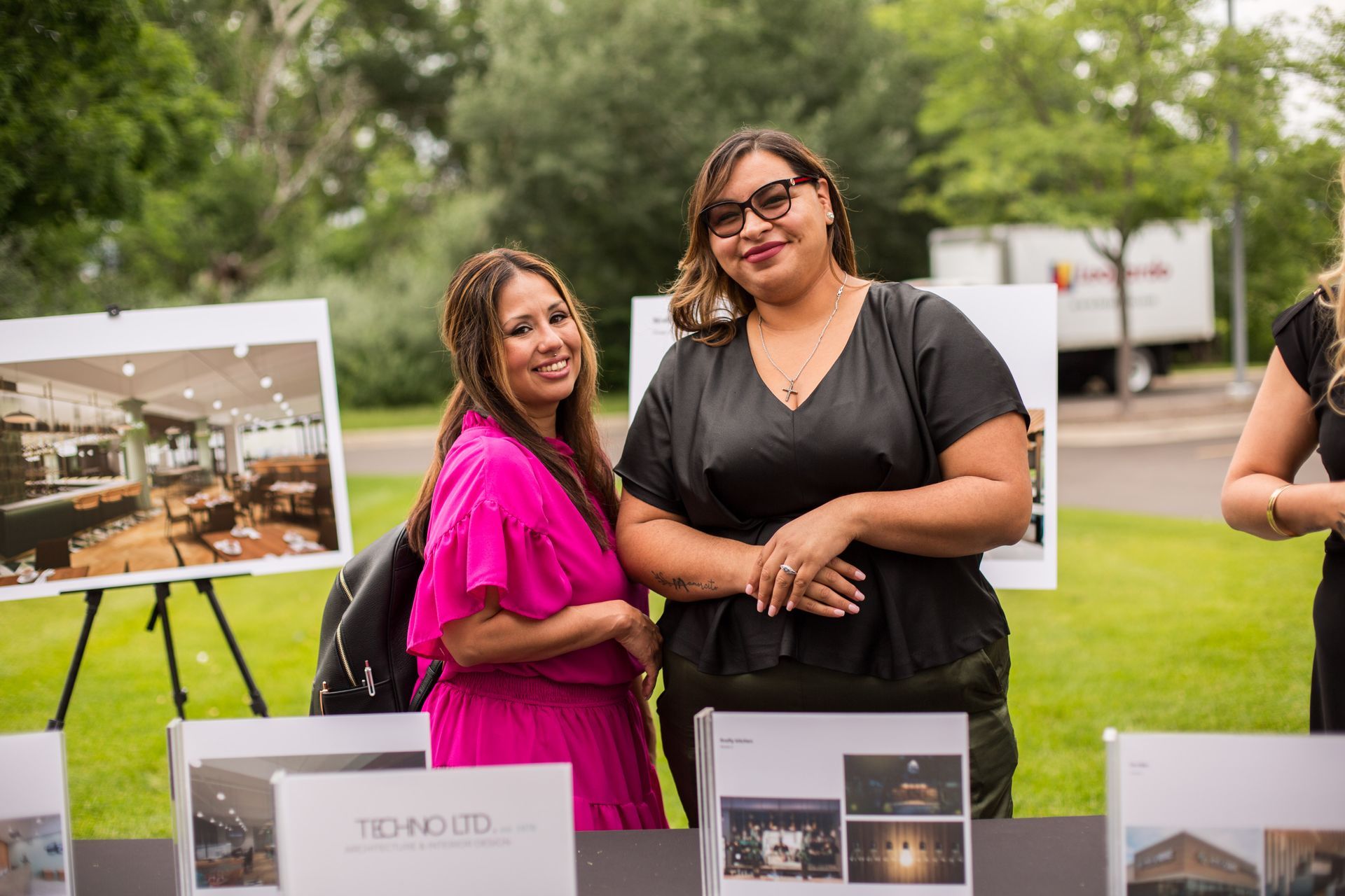 Two women smiling at an outdoor event display table with property photos on easels