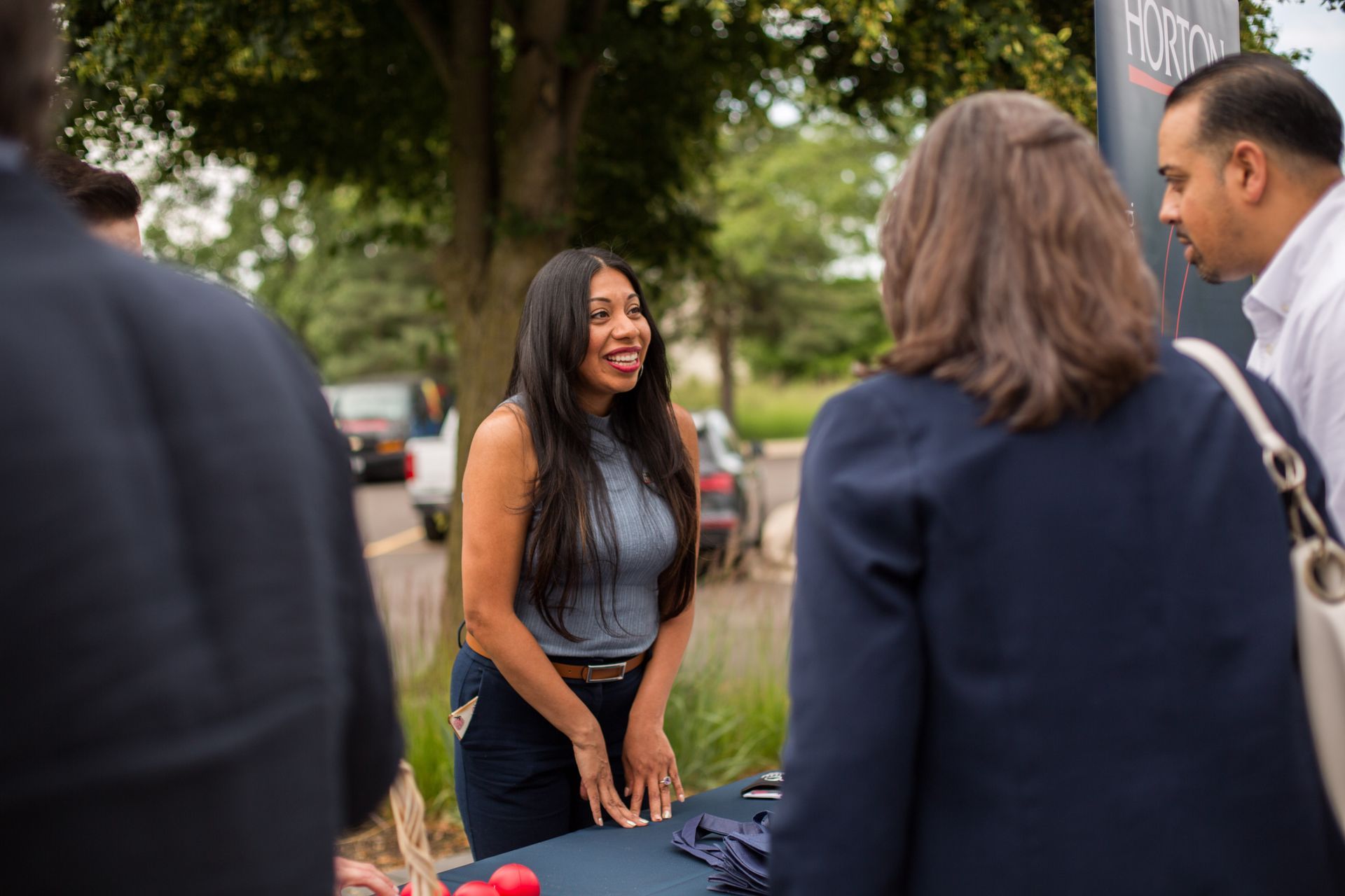 Woman speaking to a small group outdoors at a table, smiling in a park-like setting.